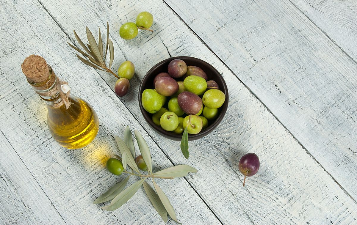 Top view Glass bottle of olive oil and wooden breakfast  bowl with raw  turkish olive seeds and leaves on white rustic vintage table. Olives background